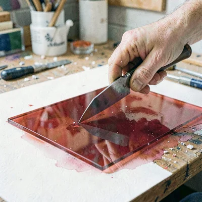 Documentary photo of a hand scoring a sheet of red glass with a steel cutter on 