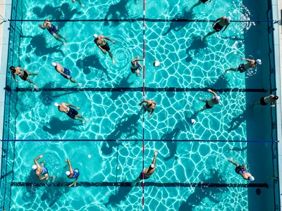 A water polo match from above, players scattered across the pool, the ball mid-f