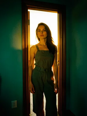 A woman standing in a doorway backlit by golden afternoon light, teal shadows on