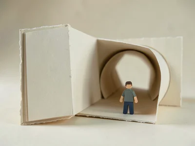 Beside a giant folded paper book opening into a tunnel, a small papercraft figur
