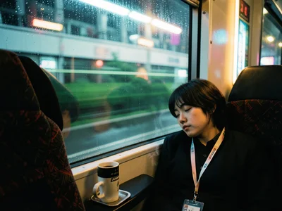 A person asleep on a commuter train with their head against the window, the morn