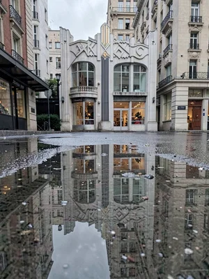 A rain puddle reflecting entire Art Deco building, real building just visible at