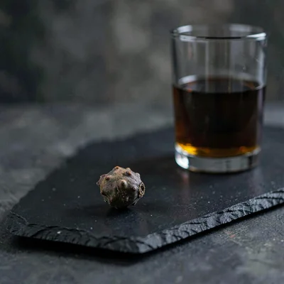 Moody still of a single juniper berry on a black slate beside a tumbler