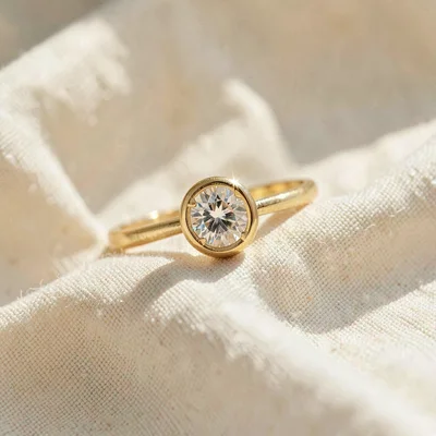 Warm product photo of a single gold solitaire ring on cream linen, soft window g