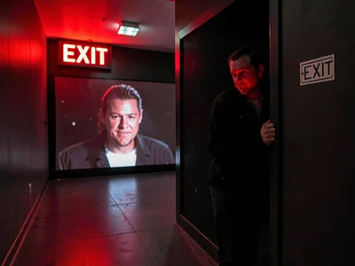 Person hiding in projection booth, movie playing beyond, EXIT sign on secret fac