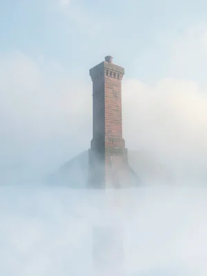 Brick chimney rising out of a fog bank, its summit catching cool light, base ent