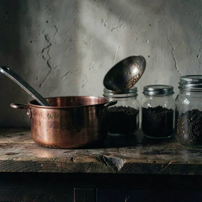 Moody scene of a wooden countertop with copper pot, ladle, and jars half in shad