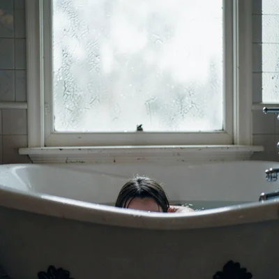 Someone in a claw-foot tub beneath a frosted window, the light diffused and pear