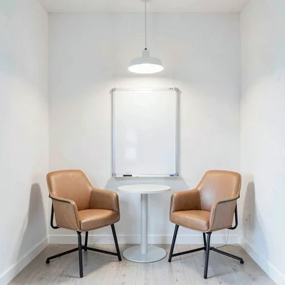 Empty whiteboard meeting nook with two tan leather chairs and a low pendant lamp