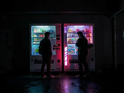 Two strangers at adjacent vending machines, the parallel commerce, acknowledged 