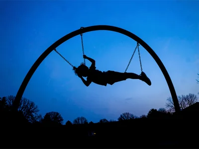 A child at apex of swing arc, body horizontal, pure black against blue twilight