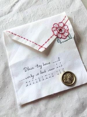An embroidered envelope on white cotton, the flap stitched closed with a red thr