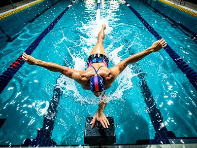 Swimmers on starting blocks mid-dive, pool stretching ahead, overhead view