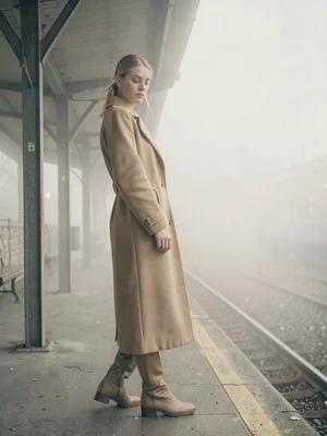 Woman in camel coat standing at a train platform, fog erasing everything beyond 