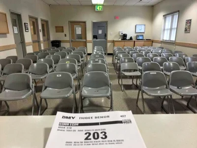 DMV waiting room, rows of plastic chairs compressed into grid, number display sh