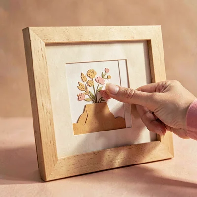 Papercraft hero of a layered paper-cut hand placing a print into a frame, warm h