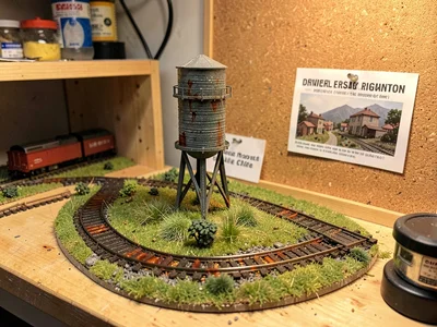 Scale model water tower beside tiny track, hand-painted rust streaks, grass made