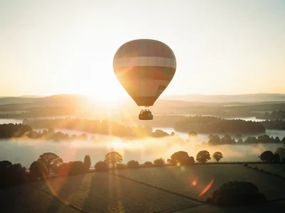 Hot air balloon floating over a valley at dawn, mist in the lowlands, warm light