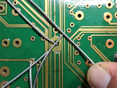 A repair technician's PCB with jumper wires bridging cut traces — silver wires o