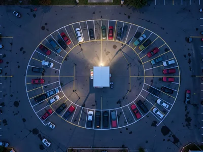 A drive-in movie theater lot from above at dusk, cars arranged in sweeping conce