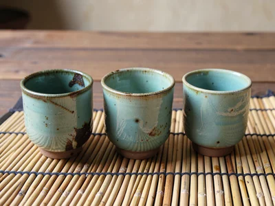 Three sake cups in a celadon glaze lined up on a bamboo mat