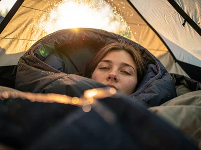 Person waking in sleeping bag, tent fabric glowing with sunrise, the disorientat