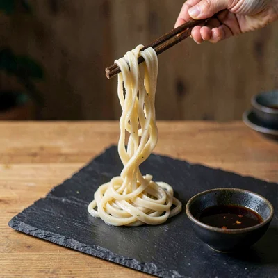 Hand-pulled fresh udon noodles dropped on dark slate, individual strands twistin