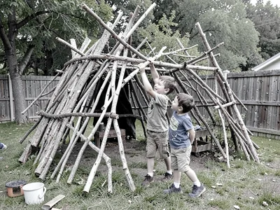 Two children building an elaborate stick fort in a backyard, one handing a branc