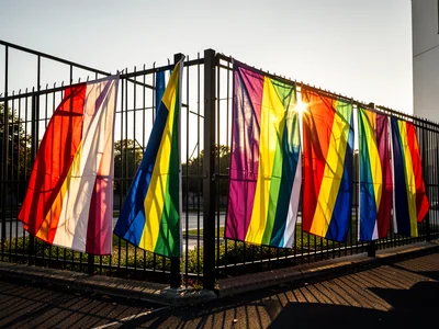 Multiple pride flags in a row along fence, breeze catching fabric, sun