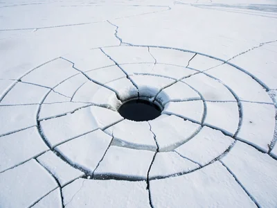 Frozen lake cracking in spiral patterns, single dark ice fishing hole punctuatin