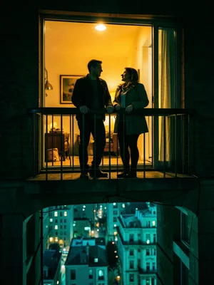 A couple on a fire escape at night, warm apartment glow behind them, the city in