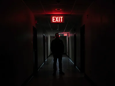 Person standing beneath glowing EXIT sign in empty hallway, 3am, the only light,