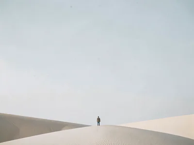 Small figure at bottom edge of endless sand dune under pale sky