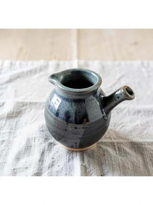 A small pouring vessel with a kinked spout and ash glaze drip, on rough linen