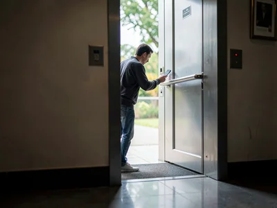 Person checking phone in elevator doorway, holding the door, the threshold betwe