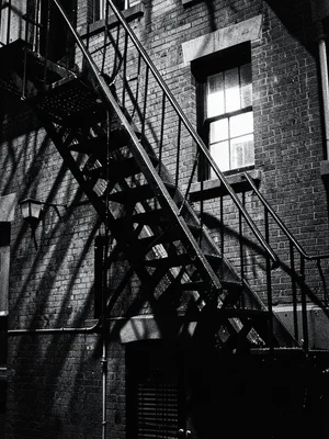 Fire escape zigzagging down a brick building, single lit window, noir shadows