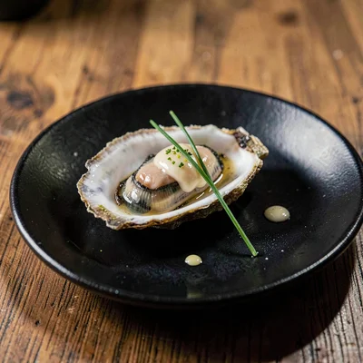 Single fresh oyster on a black ceramic plate with a tiny dot of mignonette and a