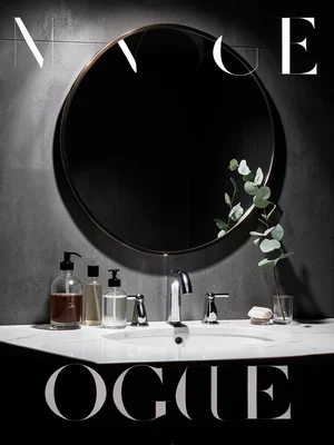 Bathroom vanity with glass bottles, a round mirror, and a single eucalyptus spri