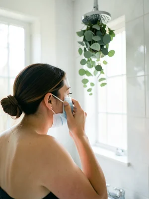 Person applying face mask in a bright bathroom, eucalyptus hanging from shower h