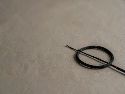 A single coil of black silk cord resting low-right on warm-grey paper, fibres ca