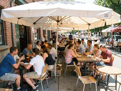 Locals crammed under single cafe umbrella at noon, empty sun-blasted tables ever
