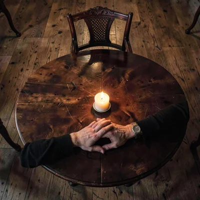 A séance table from above, hands joined in circle, one empty chair with candle