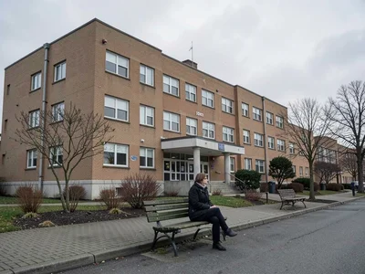 The exterior of a hospital on an overcast day, someone sitting on a bench outsid