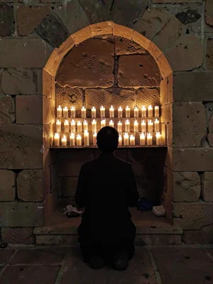 Rows of prayer candles in stone alcove, single figure kneeling in silhouette