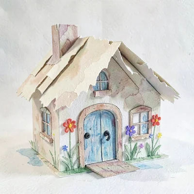 A pop-up paper cottage with torn-paper thatched roof and crayon flowers at the d