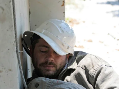 A construction worker asleep in a sliver of shade during midday break, hard hat 