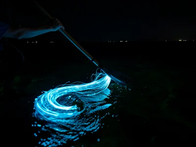 Kayak paddle pulling through dark water — a spiral of bright blue fire trailing 