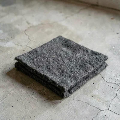 One folded square of charcoal wool felt arranged neatly on washed concrete, fibe