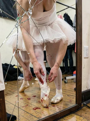 A ballet dancer removing pointe shoes backstage, feet bloodied, reflection in cr
