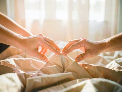 Two hands reaching toward each other across rumpled linen sheets, morning light 